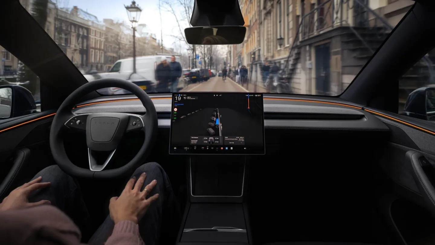 Tesla’nın kabininden, FSD sürüş ekranı ve elleri direksiyon yakınında görünen sürücüyle şehir içi yol görüntüsü.
