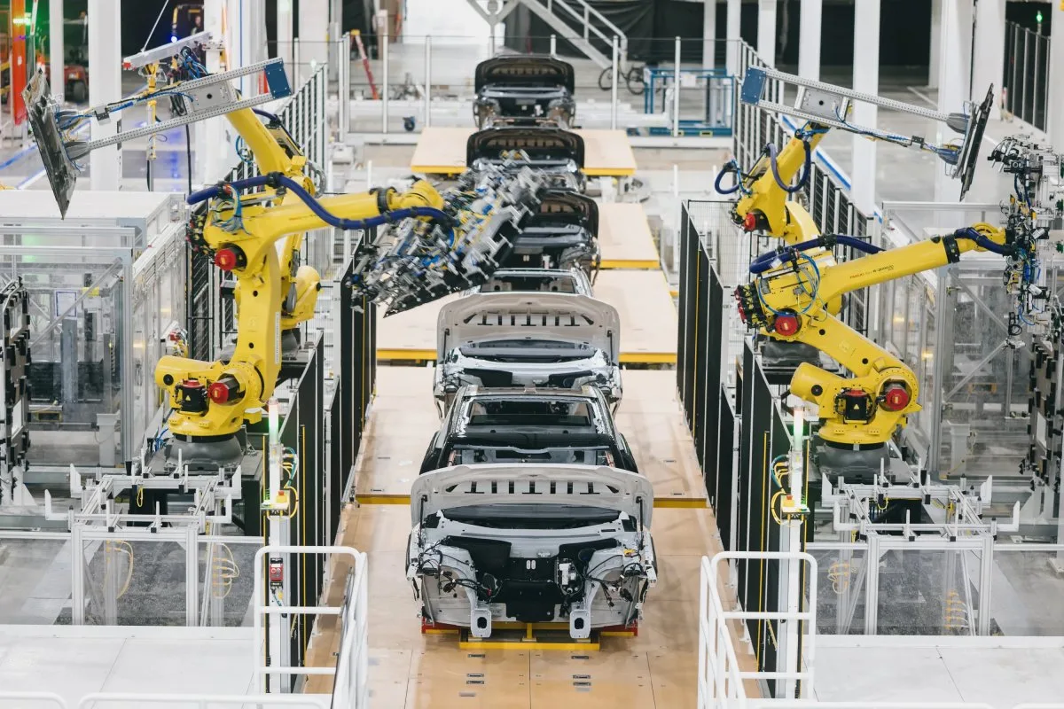 Bir otomobil fabrikasında sarı robot kollar araç gövdeleri üzerinde çalışıyor.