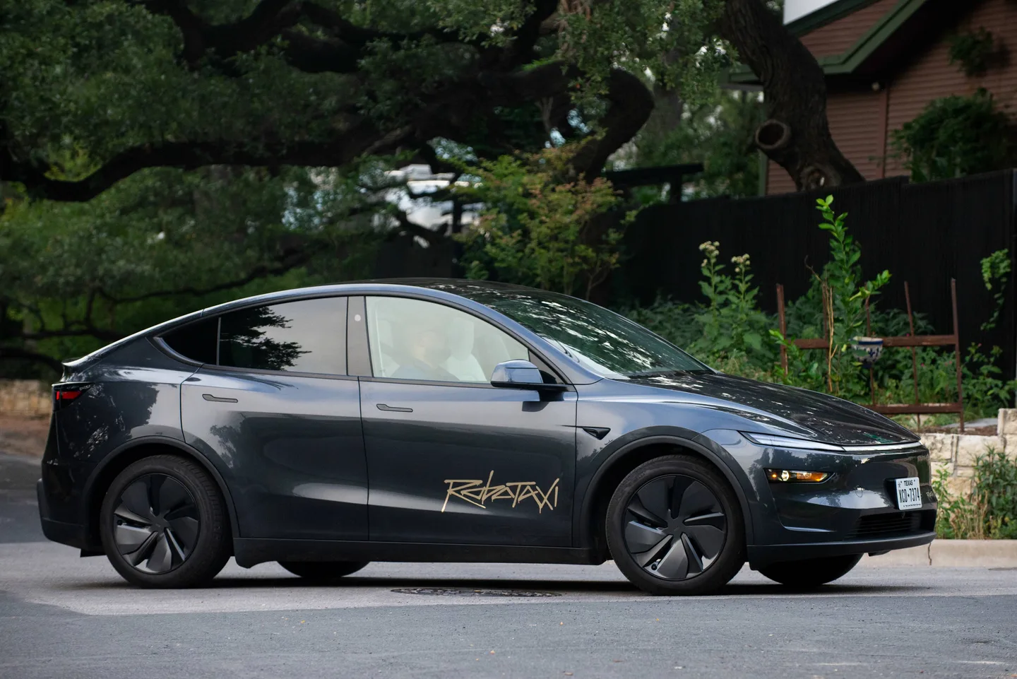 Yan kapısında “Robotaxi” yazısı bulunan koyu renkli Tesla Model Y, ağaçların önünde park hâlinde.